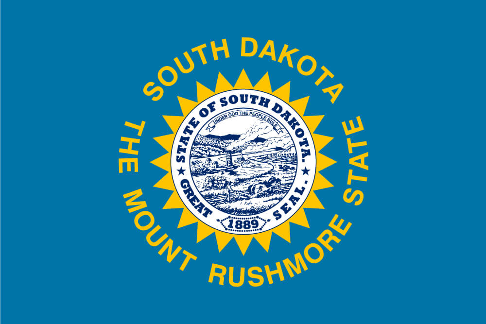 a field of sky blue charged with the state seal in blue and white and surrounded by a serrated, golden sun and the words ”South Dakota” and “The Mount Rushmore State”